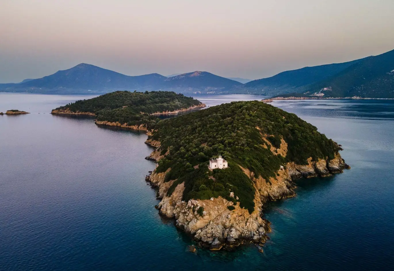 Aerial view of a forested peninsula at twilight, featuring a small white building near the rocky tip, set against distant mountains and calm blue waters.