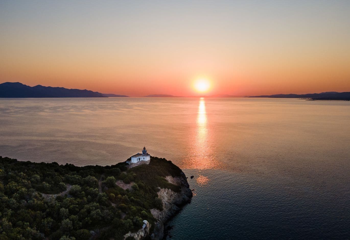 Aerial sunset view of a white lighthouse on the tip of a green peninsula overlooking calm waters.