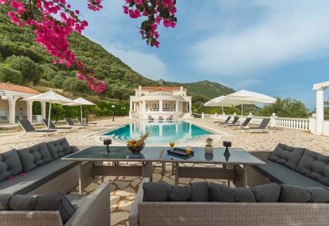 Luxury villa outdoor patio with swimming pool, sun loungers, outdoor dining set, pink bougainvillea, and green hill backdrop