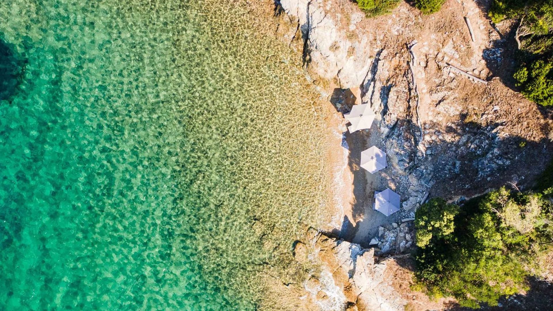 Aerial view of a secluded cove with clear, shallow turquoise water washing onto a rocky shore dotted with a few sun umbrellas.