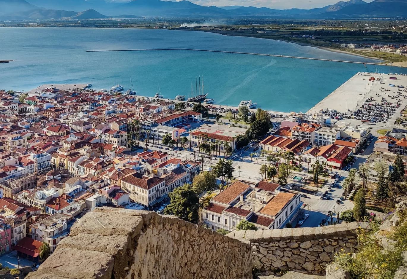 Aerial view of the coastal town of Nafplio, featuring red-tiled roofs, a busy harbor, and distant mountains across the bay.