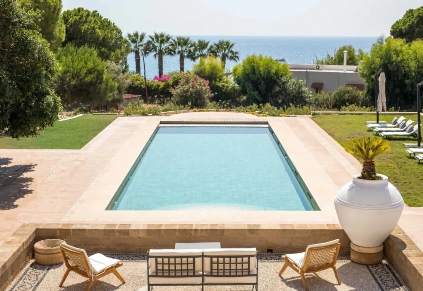 Rectangular outdoor swimming pool surrounded by a patio, lush garden, and sea view in the background