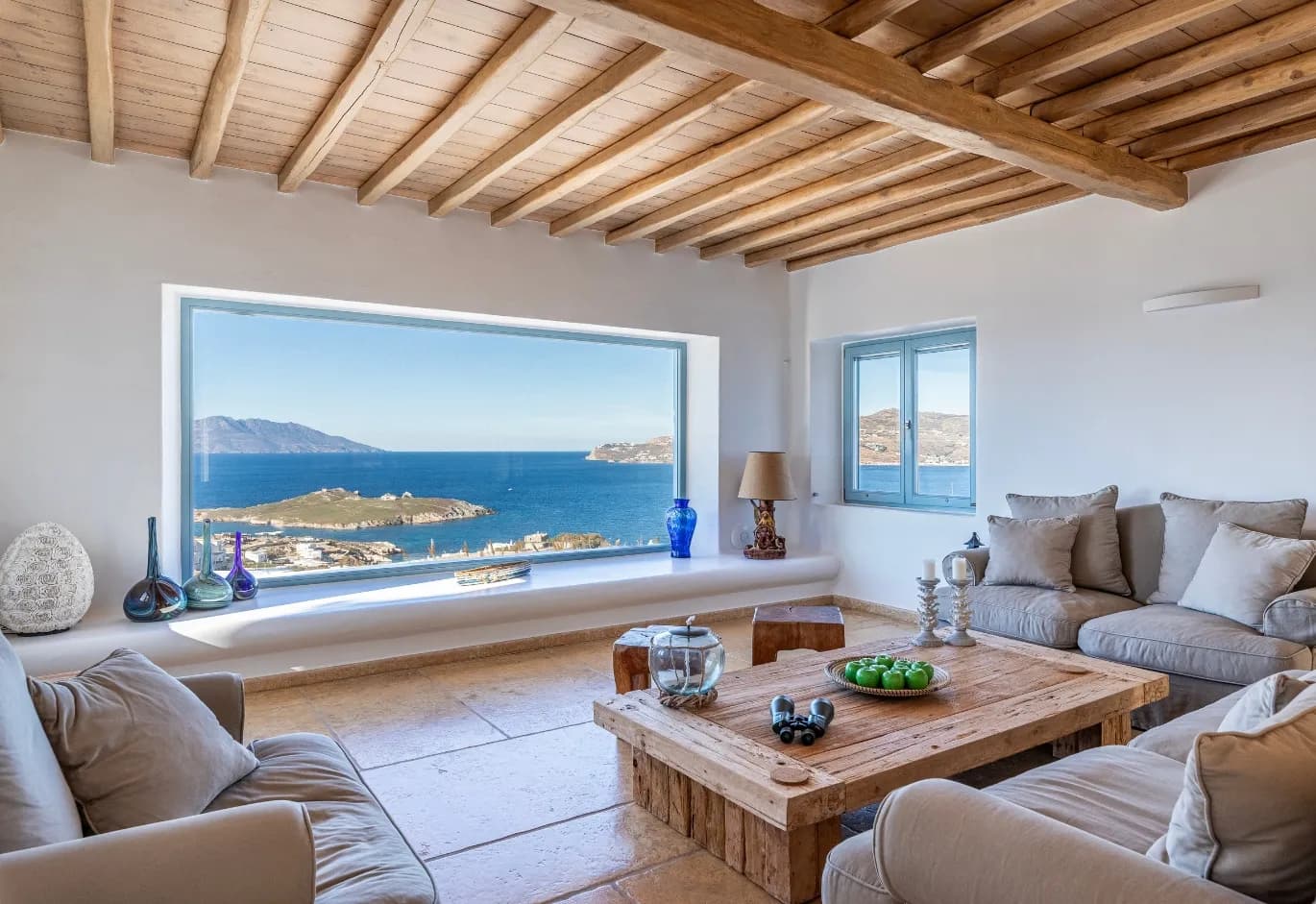Cycladic-style living room with exposed wooden beams, rustic coffee table, neutral sofas, and a large picture window overlooking the Aegean sea