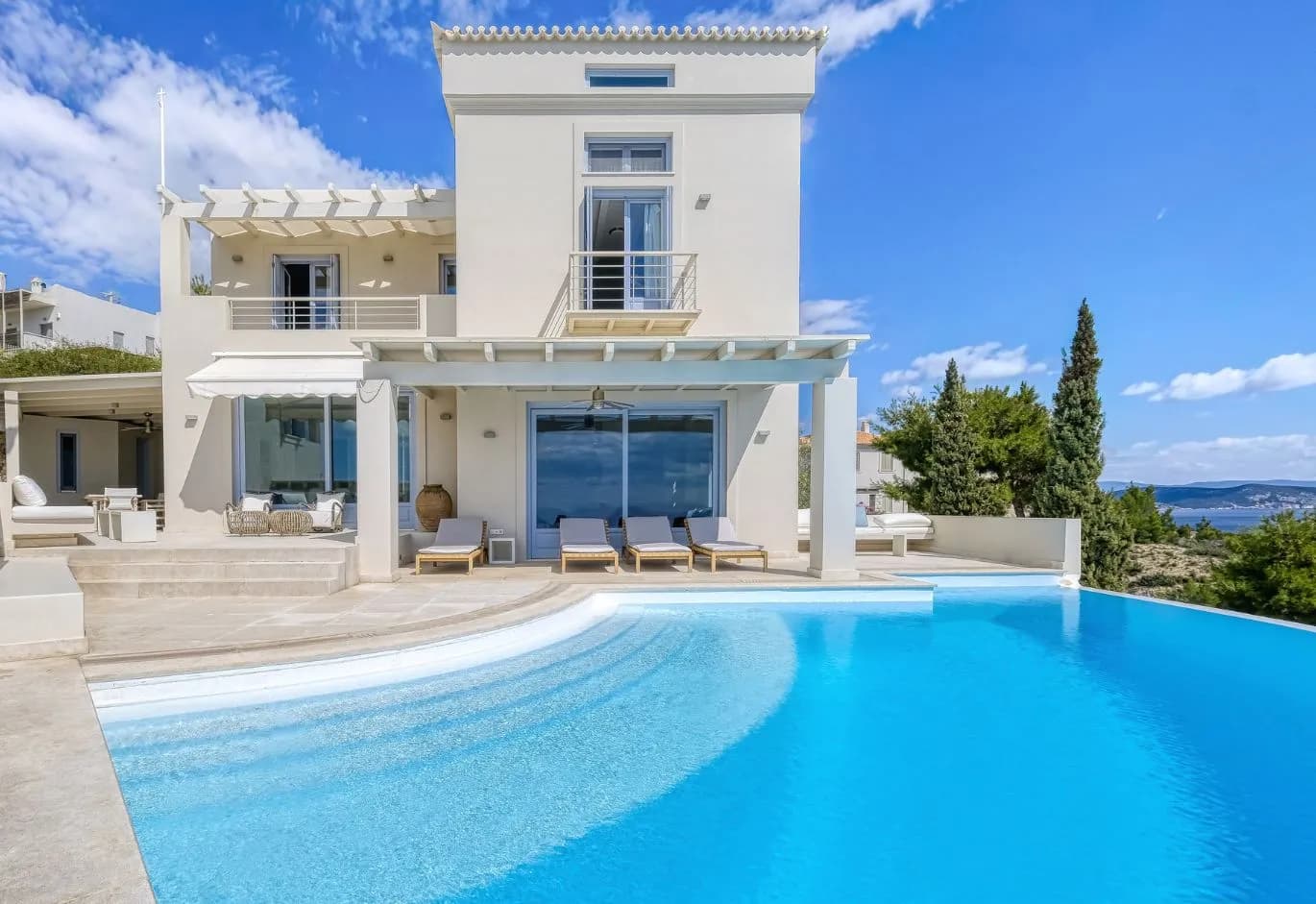 Modern luxury villa with a curved infinity pool, shaded sun loungers, and expansive glass doors facing the sea.