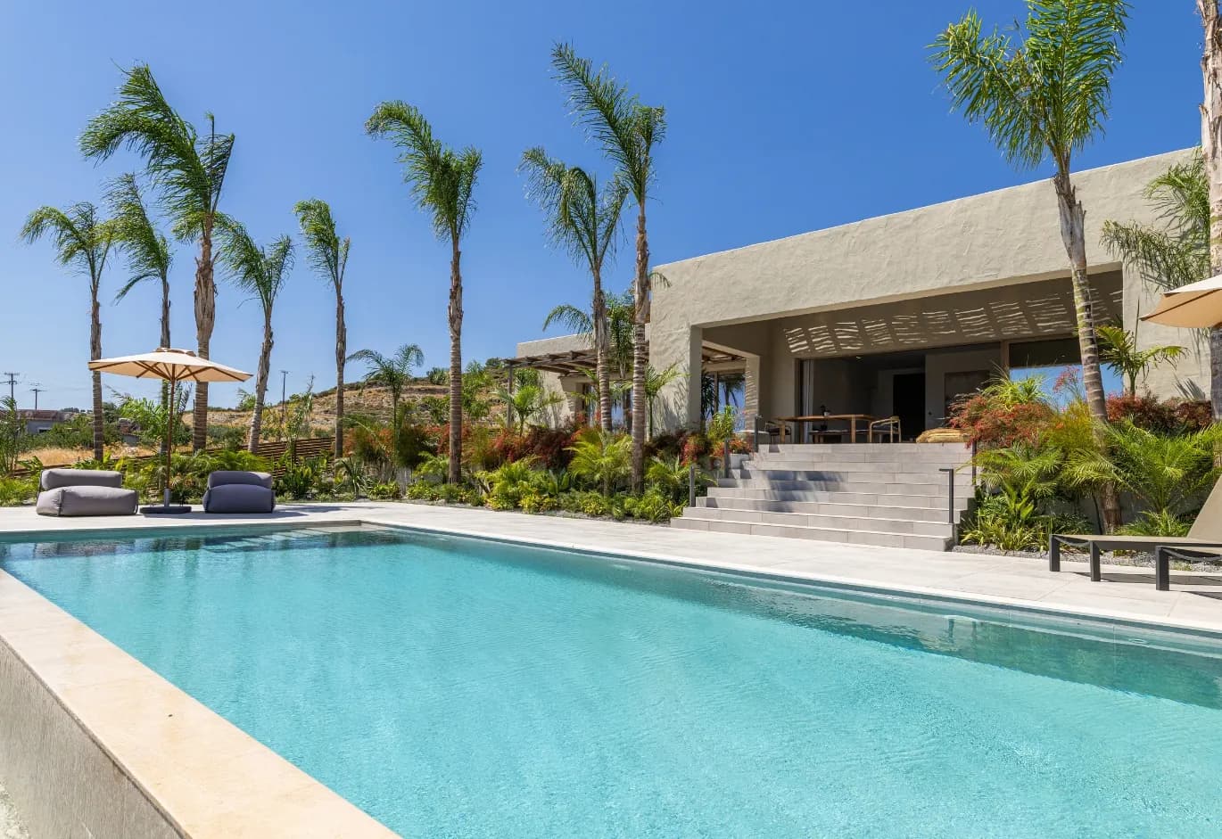 Modern villa patio with clear blue pool, palm trees, sun beds, and limestone steps leading to the main house
