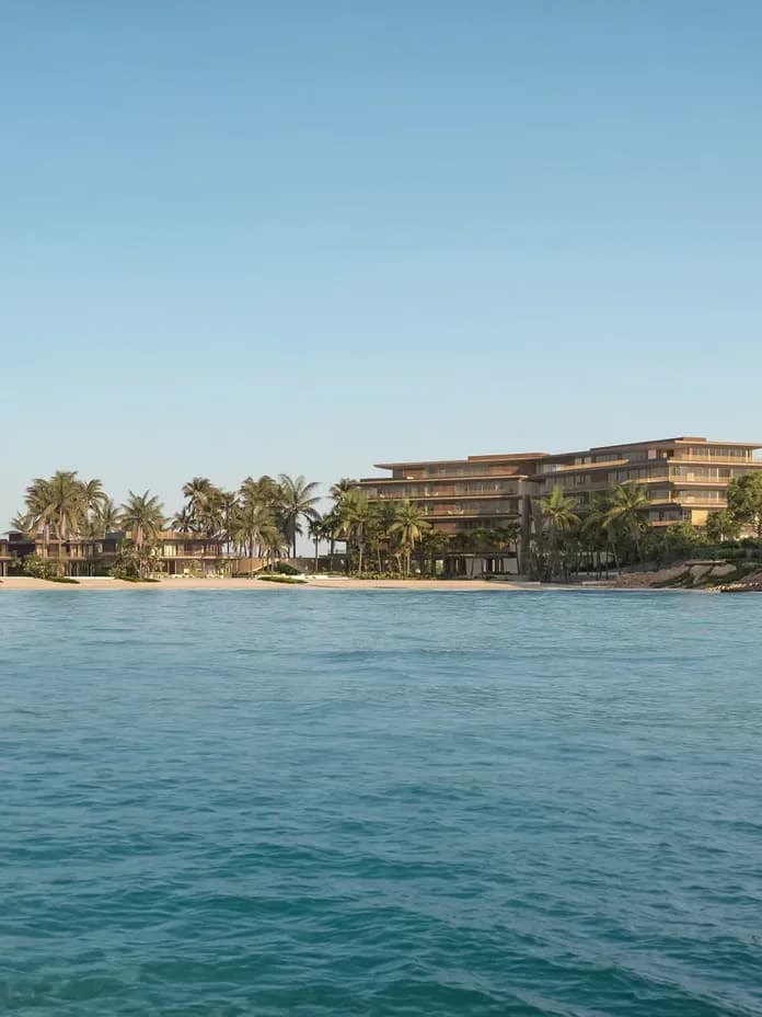 Beachfront resort with palm trees, sandy shore, and modern buildings under a clear blue sky, viewed from the ocean.
