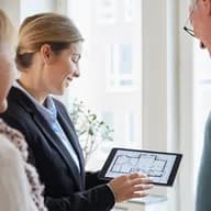 A real estate agent shows a floor plan on a tablet to a senior couple. The agent is smiling.