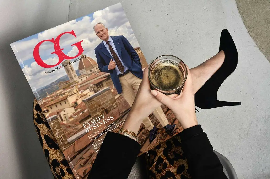 Person holding a magazine featuring a man in a suit, with a cityscape background, and a glass of coffee, wearing black high heels and leopard print pants.