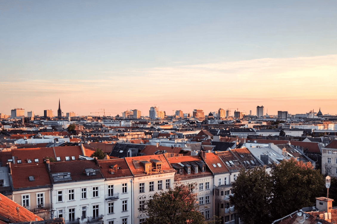 Aerial view of a cityscape at sunset with red-roofed buildings in the foreground and tall modern structures in the background under a clear sky.