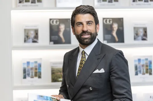 Smiling man in a dark suit with a patterned tie and pocket square, holding a brochure, standing in front of shelves filled with magazines.