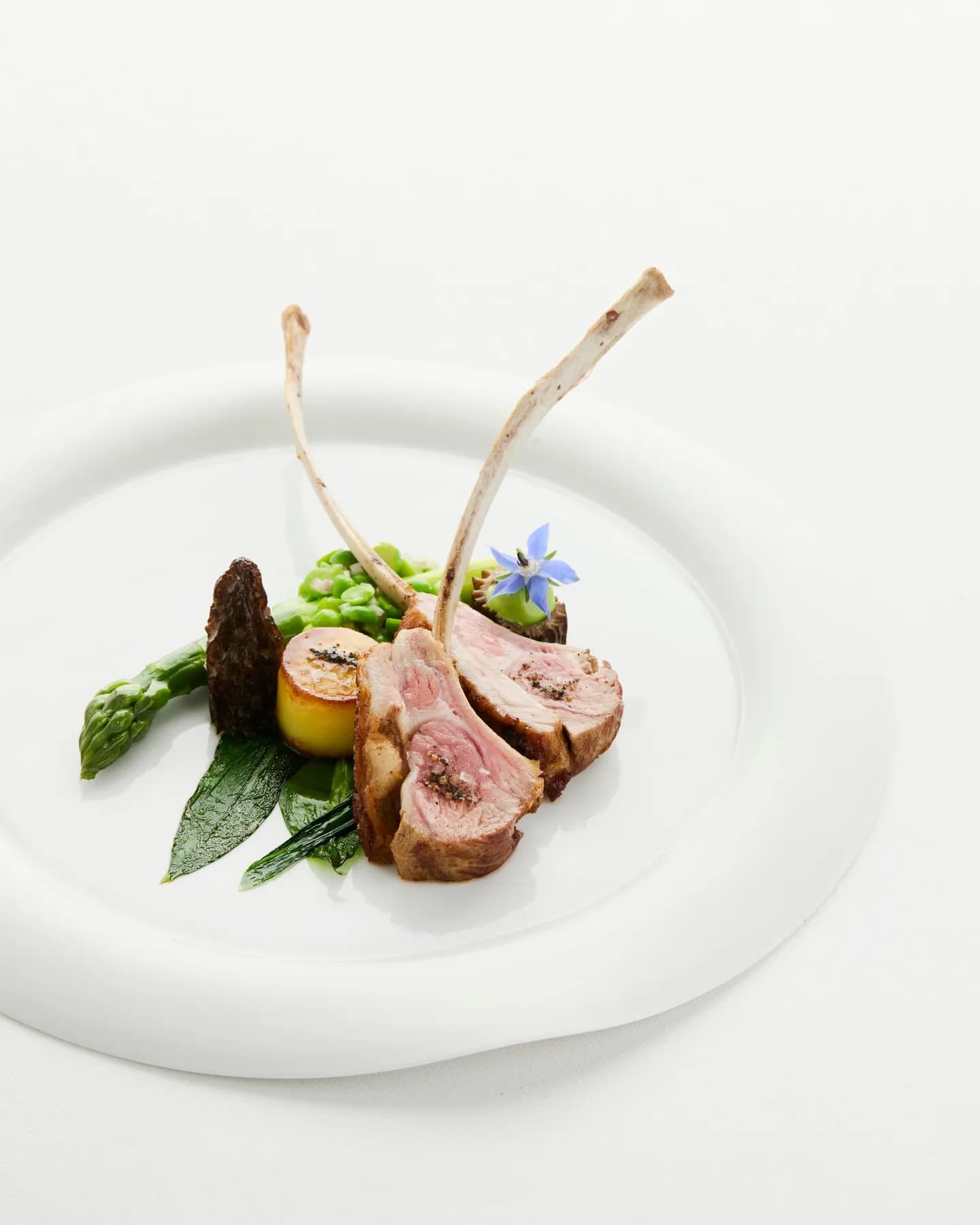 Elegant lamb rack with two upright bones, sliced lamb, asparagus, peas, a roasted potato, greens, and a blue flower garnish.