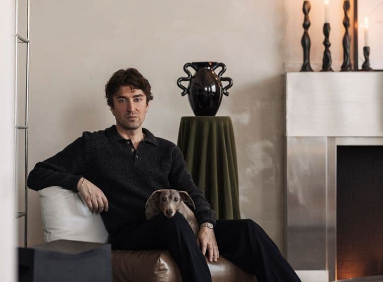 Man seated on a brown cushion, with a small dog in his lap, in a modern living room with a decorated mantle and candlesticks.