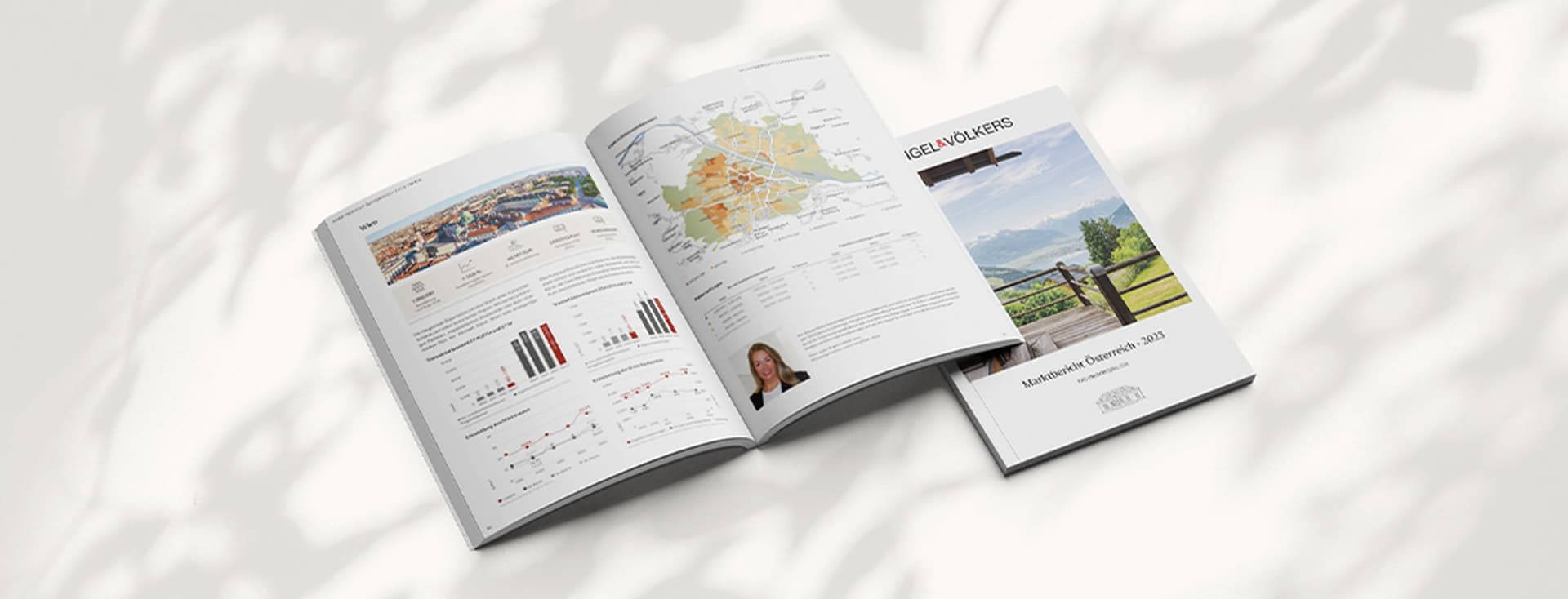 Aufgeschlagenes Marktbericht-Heft mit Diagrammen, Karten und Texten, daneben das Deckblatt mit Landschaftsfoto und der Aufschrift „Marktbericht Österreich 2023“ von Engel & Völkers, auf hellem Untergrund mit weichen Schattenspielen.