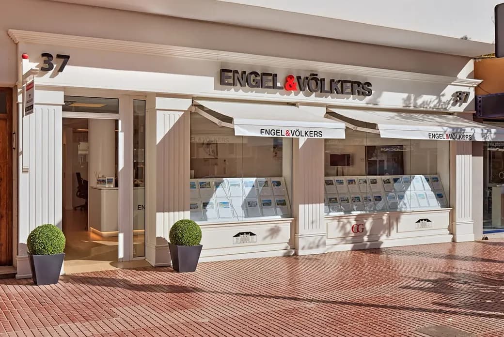 Elegant white Engel & Völkers real estate storefront with awnings, window property listings, two potted topiary plants and brick sidewalk