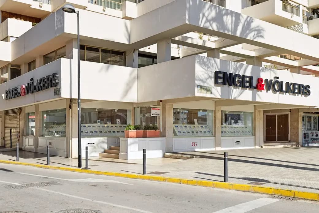 Sunlit corner Engel & Völkers real estate storefront, white facade with large sign, display windows of property listings and potted plants.