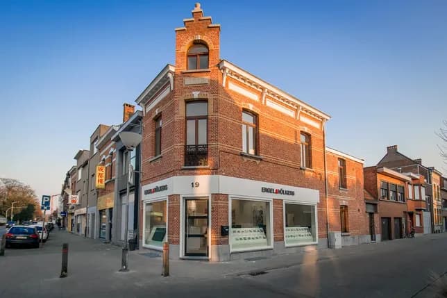 A corner brick building with large windows, housing Engel & Völkers, surrounded by parked cars and adjacent buildings under a clear sky.