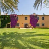 Casa amarilla de dos pisos con buganvillas moradas, hiedra verde y palmeras en un día soleado.