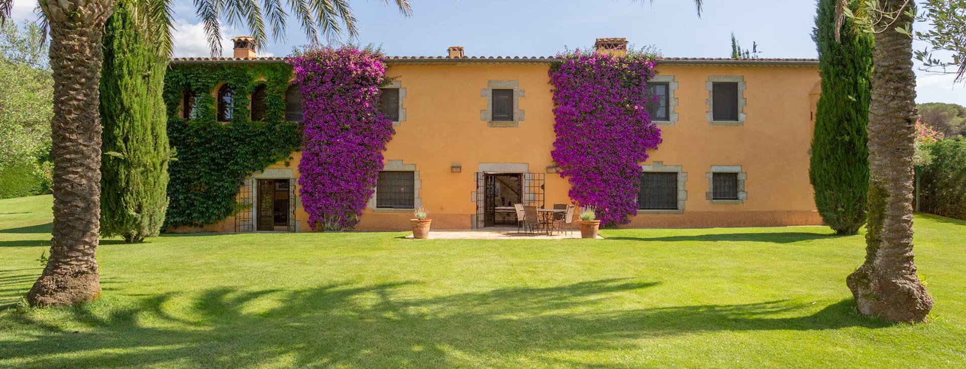 Zweistöckiges gelbes Haus mit lila Bougainvillea, grünem Efeu und Palmen an einem sonnigen Tag.