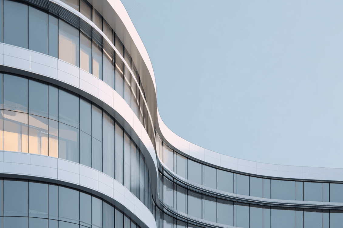 Curved modern glass office building with white horizontal bands and one warm-lit window against pale blue sky
