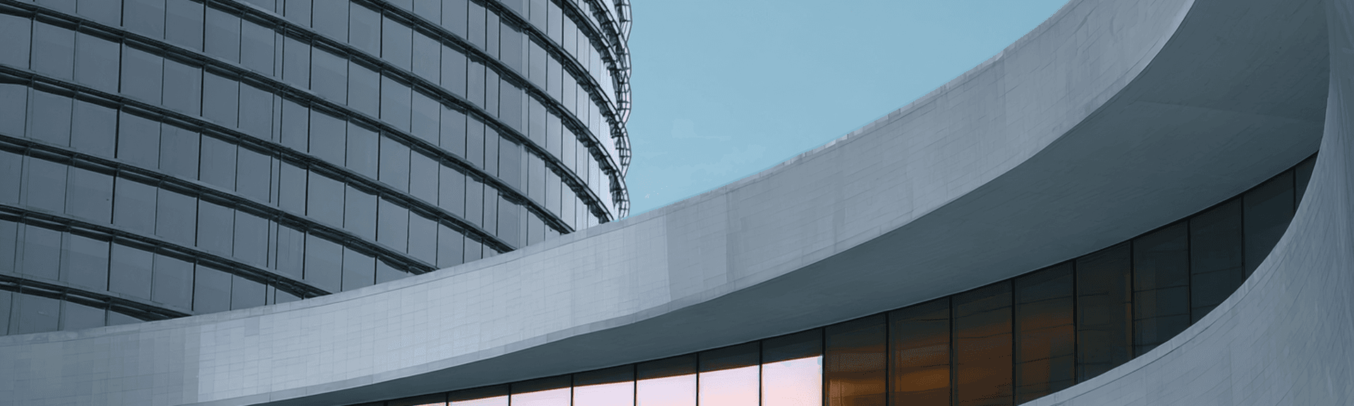 Modern building with curved concrete facade, glass windows reflecting sunlight, and adjacent sleek glass skyscraper under a clear blue sky.