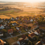 Aerial view of a small village with red-roof houses, green lawns and patchwork farmland bathed in golden sunset.