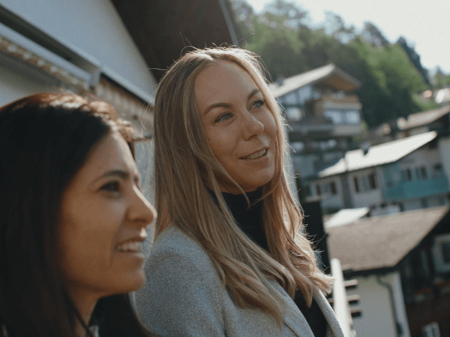 Two women stand outdoors in a hillside village, smiling, with houses and trees in the background.