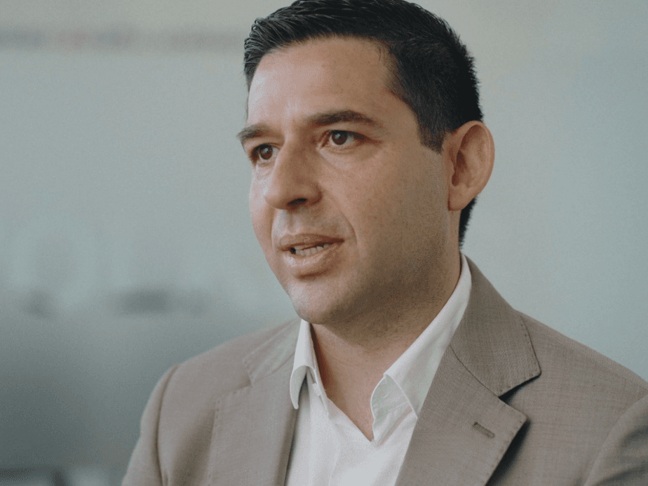 A man in a beige suit and white shirt stands in a bright office, looking to the left.