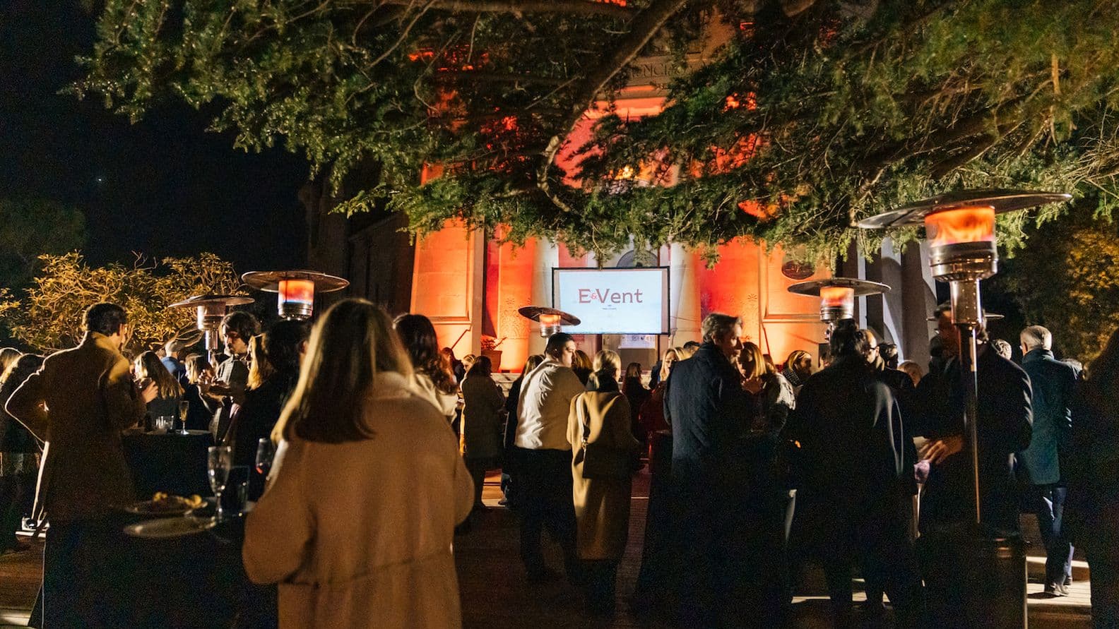 Outdoor evening event with a crowd of people, patio heaters, and a screen displaying "E&Vent" in front of a building lit with orange lights.