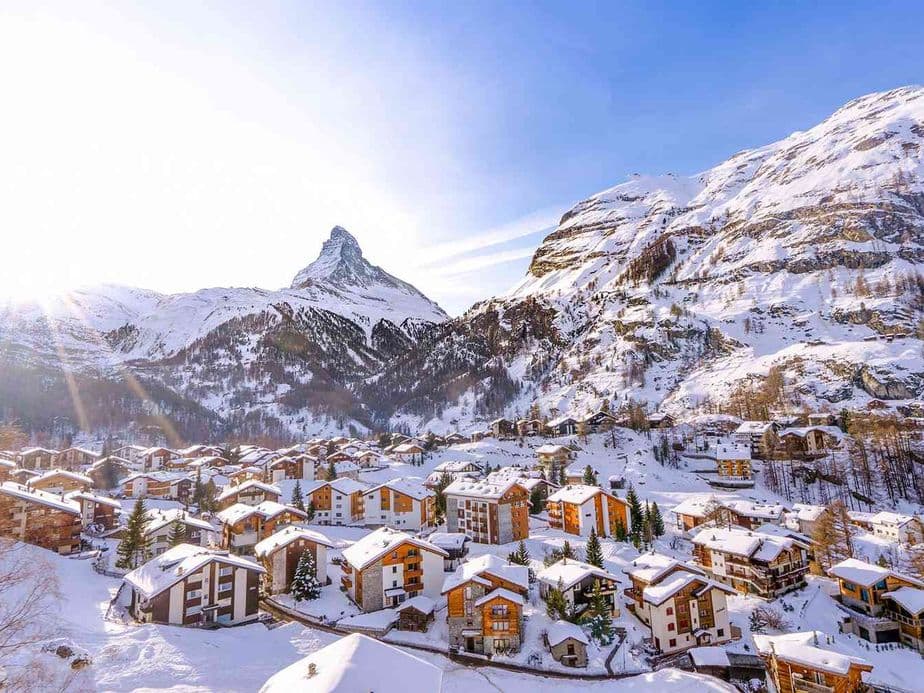 Snow-covered alpine village with wooden chalets, surrounded by mountains under a clear blue sky, and the Matterhorn in the background.