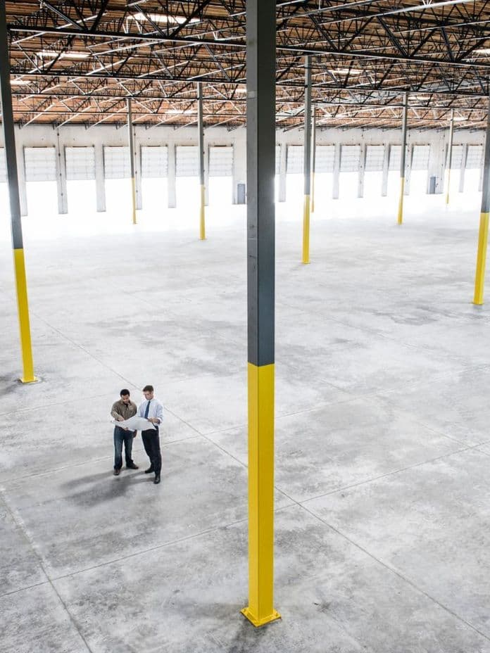Two people stand in a large, empty warehouse with high ceilings, yellow pillars, and numerous loading doors in the background.