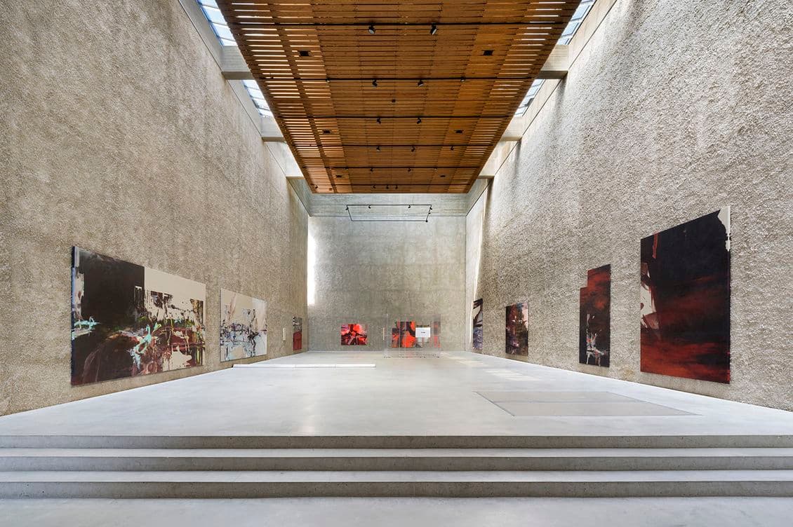 A spacious art gallery with high ceilings, textured walls, and large abstract paintings displayed on either side of the room.