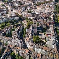 Luftaufnahme eines dicht bebauten Stadtgebiets mit einer Kirche, umgeben von Wohnhäusern und von Bäumen gesäumten Straßen