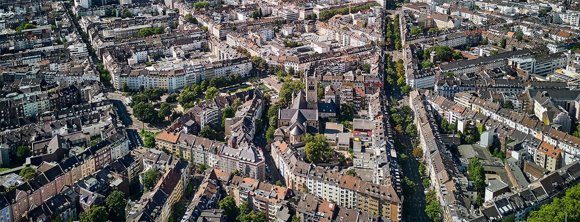 Luftaufnahme eines dicht bebauten Stadtgebiets mit einer Kirche, umgeben von Wohnhäusern und von Bäumen gesäumten Straßen
