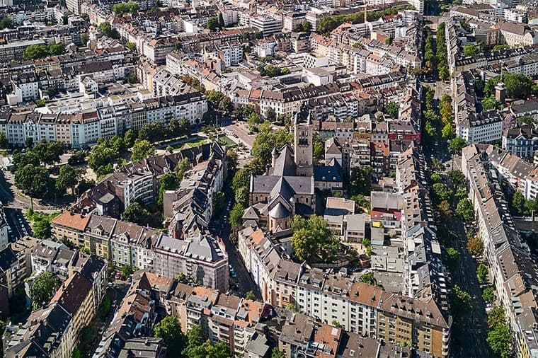 Luftaufnahme eines dicht bebauten Stadtgebiets mit einer Kirche, umgeben von Wohnhäusern und von Bäumen gesäumten Straßen