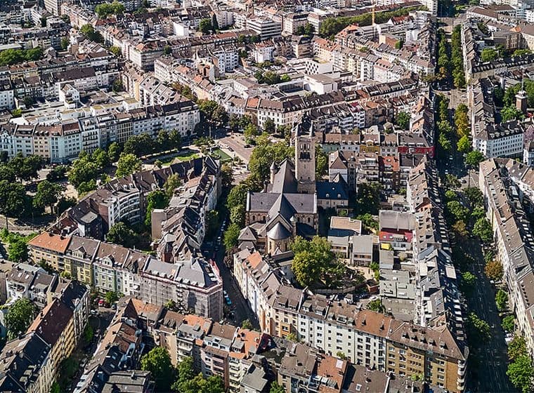 Luftaufnahme eines dicht bebauten Stadtgebiets mit einer Kirche, umgeben von Wohnhäusern und von Bäumen gesäumten Straßen