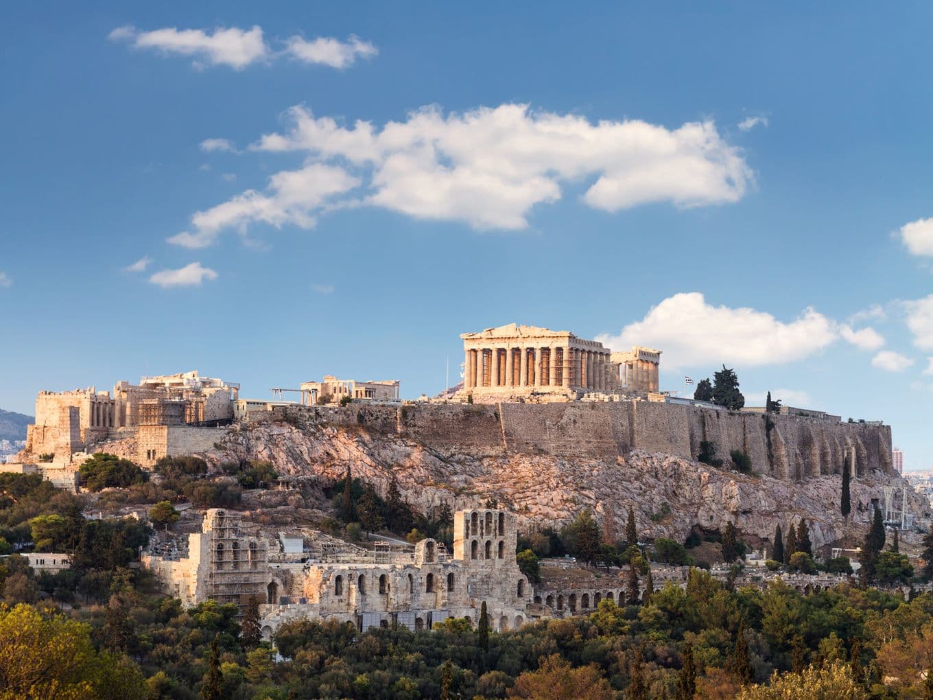The Acropolis in Athens, Greece, with the Parthenon atop a rocky hill, under a blue sky with scattered clouds. Surrounding greenery visible.