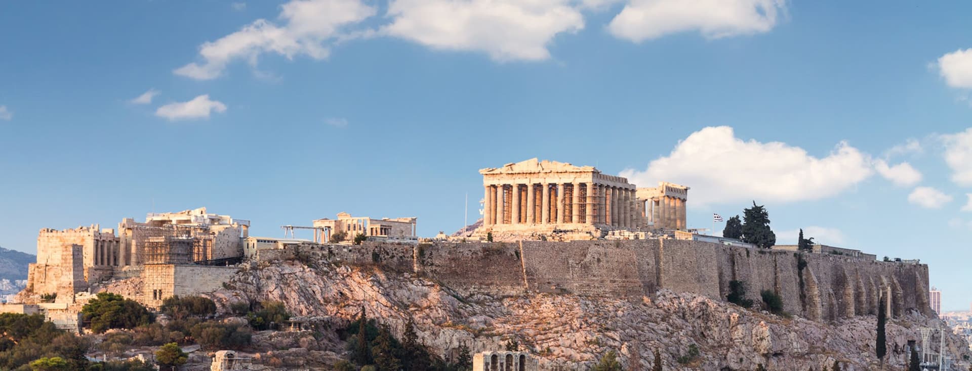 The Acropolis in Athens, Greece, with the Parthenon atop a rocky hill, under a blue sky with scattered clouds. Surrounding greenery visible.