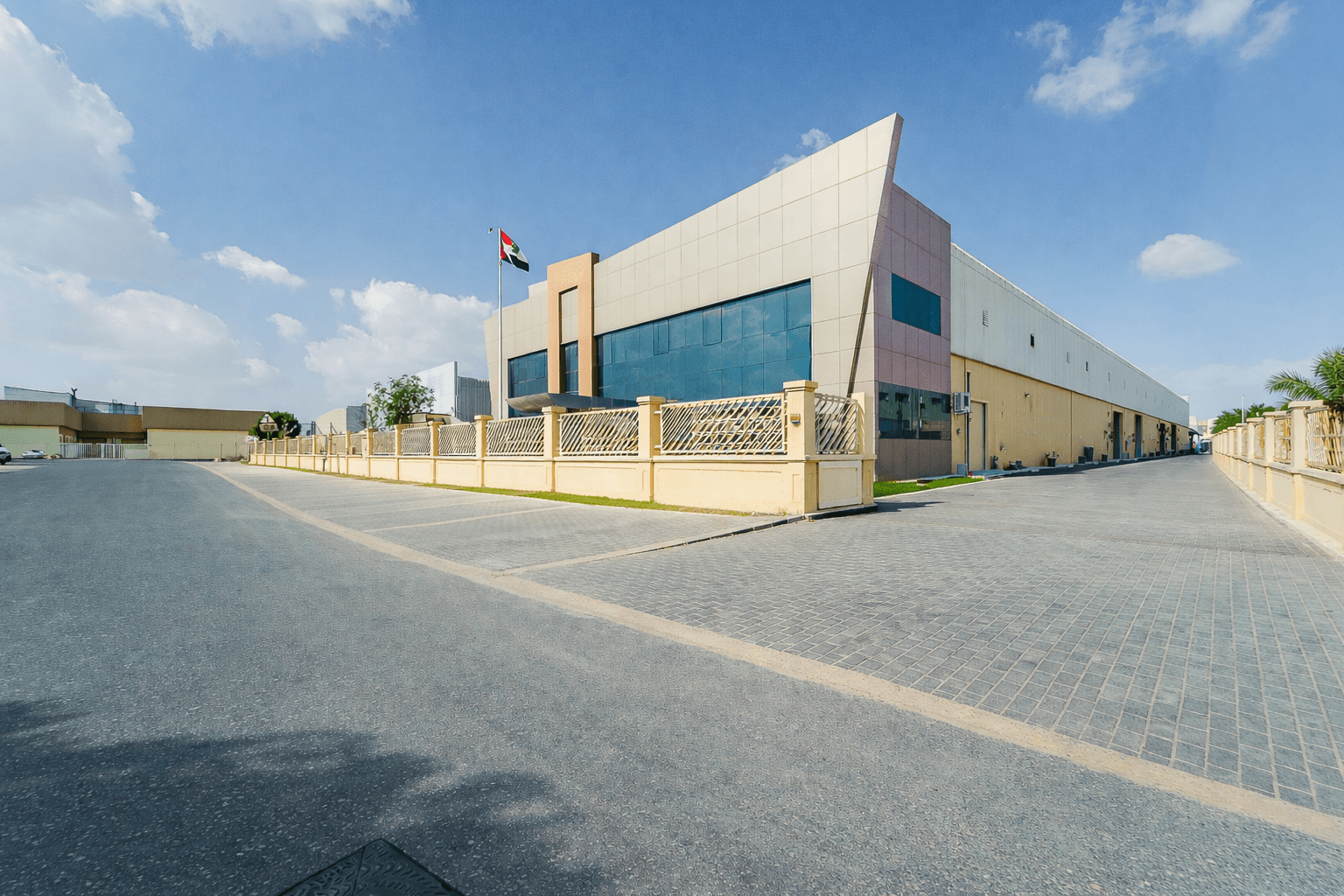 Modern industrial building with UAE flag, beige and pink facade, large windows, fenced perimeter, under a clear blue sky with scattered clouds.