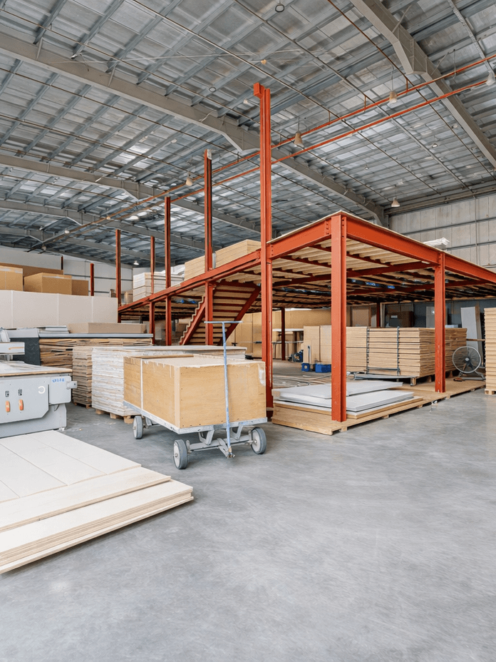 Spacious warehouse interior with stacks of wooden boards, metal shelves, and industrial equipment on a polished concrete floor.