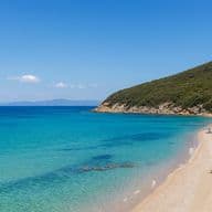 A scenic beach with turquoise water, sandy shore, and green hills. Sunbathers relax under blue umbrellas along the coastline. Clear blue sky above.
