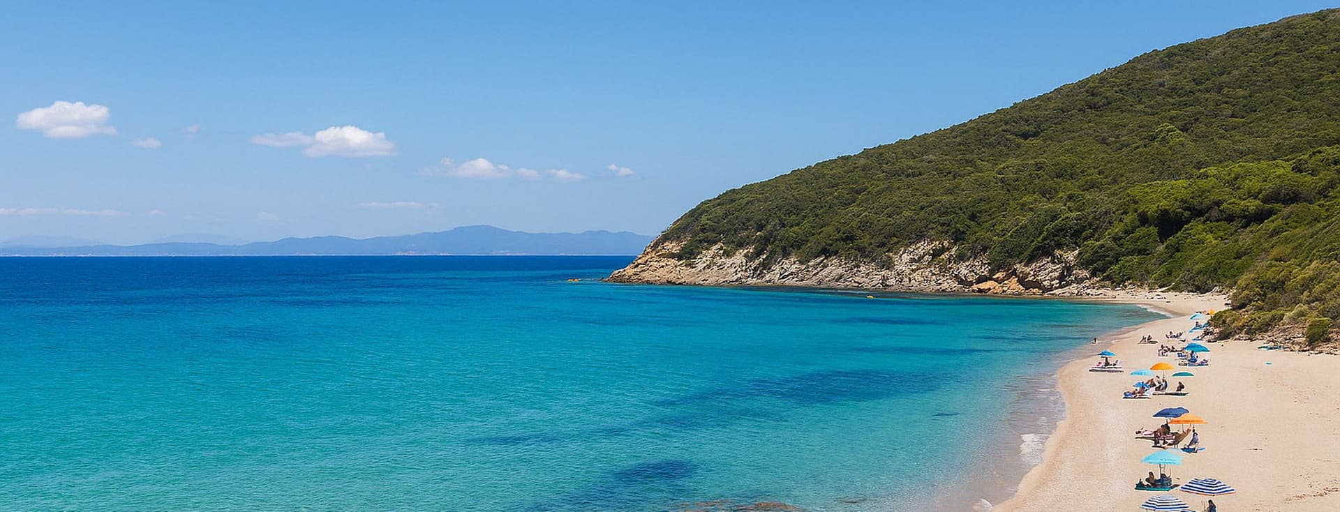 Ein malerischer Strand mit türkisfarbenem Wasser, Sandstrand und grünen Hügeln. Sonnenanbeter entspannen sich unter blauen Sonnenschirmen an der Küste. Klarer blauer Himmel darüber.