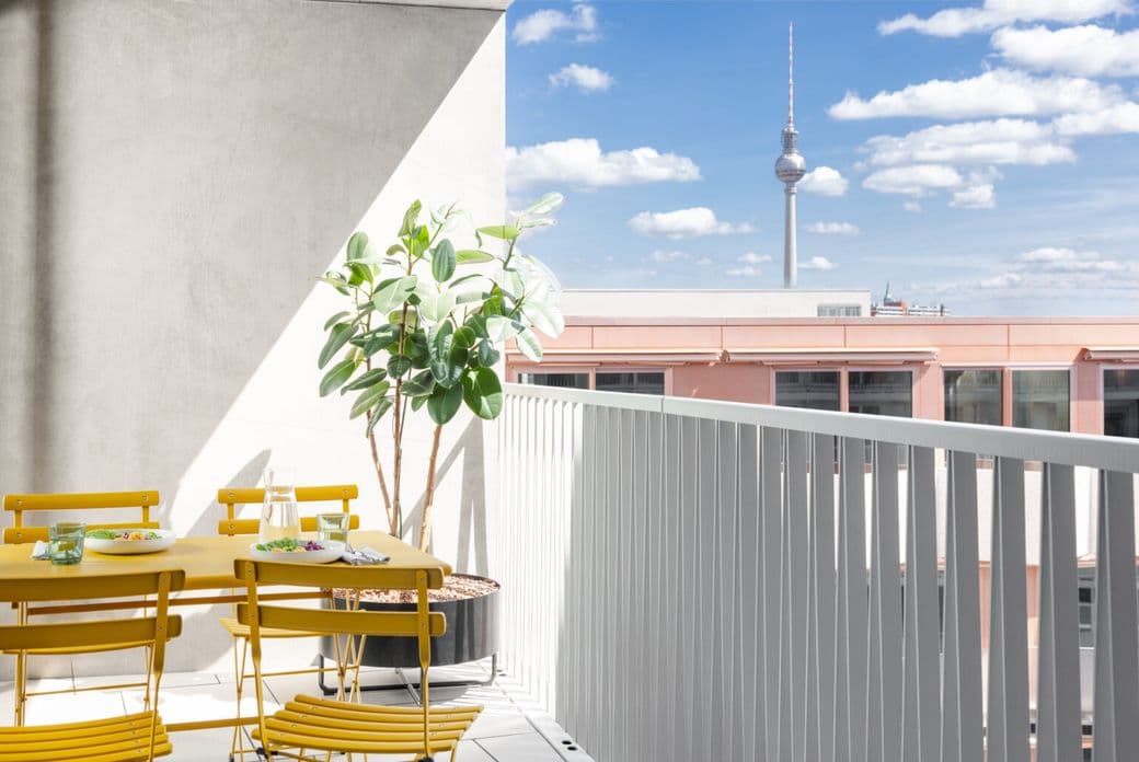 A sunny balcony with yellow chairs and table, a potted plant, and a view of a city skyline with a tower against a blue, cloudy sky.