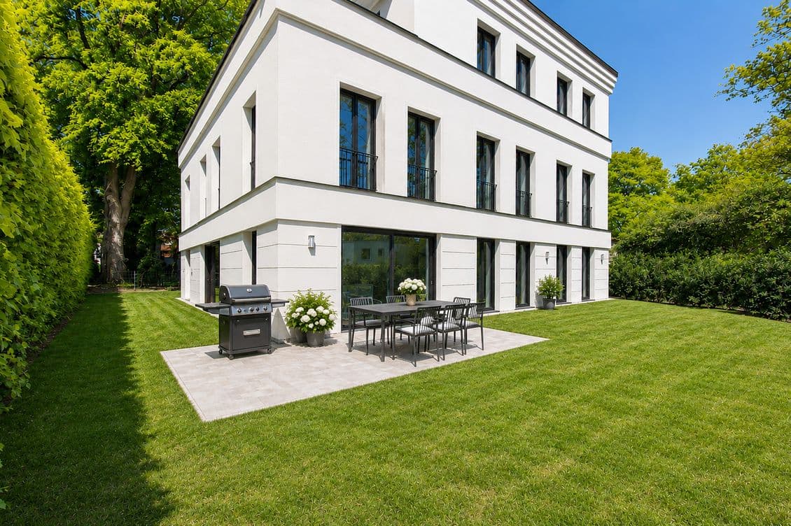 Modernes, weißes, dreistöckiges Haus mit Terrasse, Esstisch im Freien und Grill auf einem gepflegten Rasen, flankiert von Hecken und hohen, belaubten Bäumen.