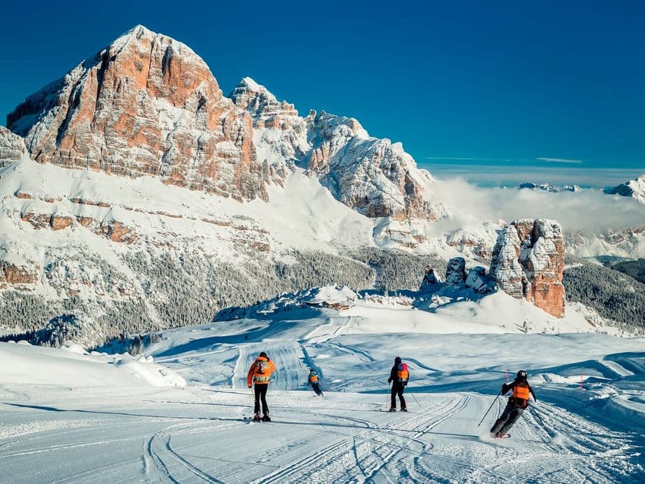 Skiers descending a snowy slope with majestic, snow-covered mountains in the background under a clear blue sky.