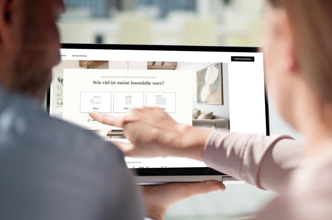 Two people using a tablet to view a real estate website with a question in German about property value. One person points at the screen.