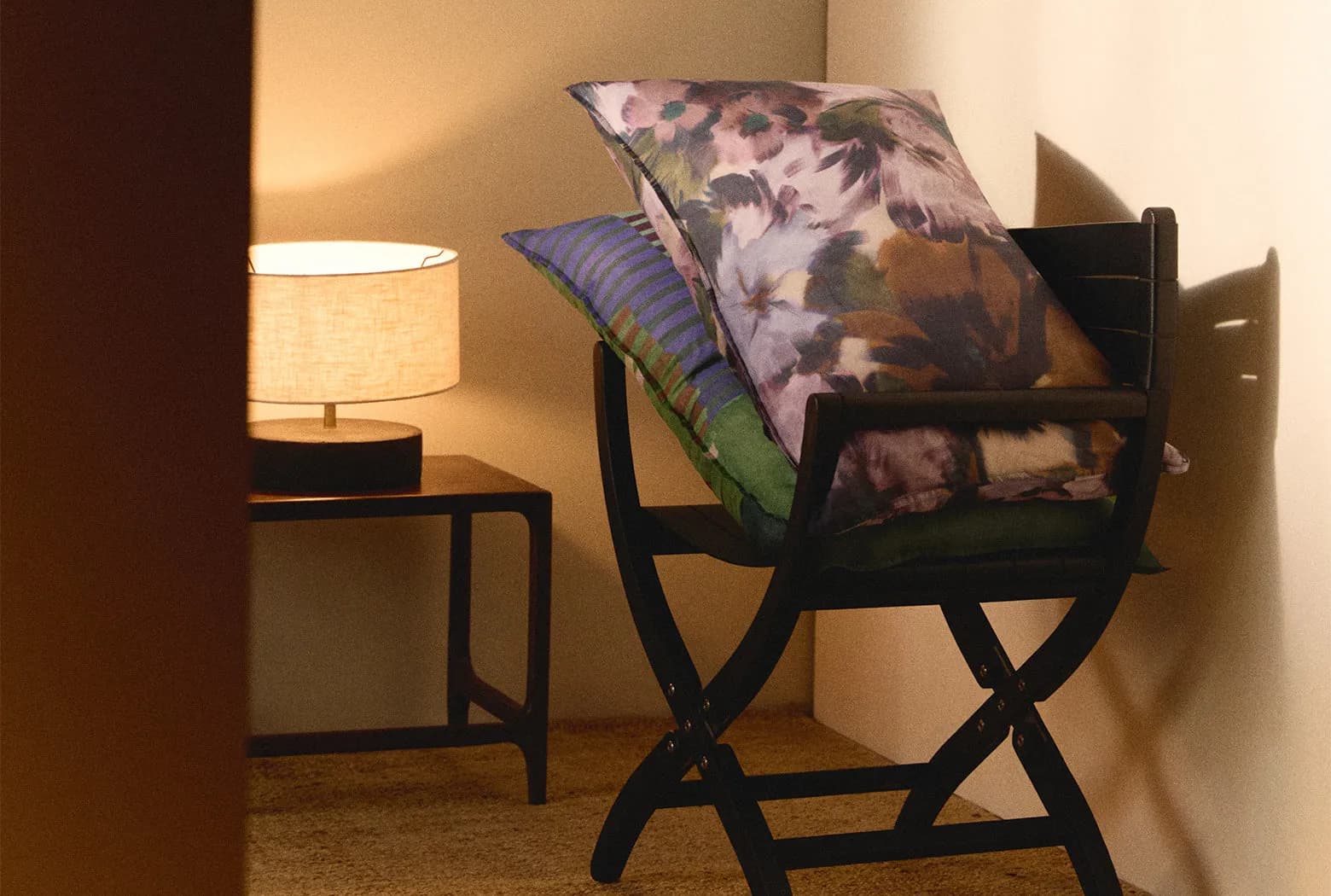 A dark wooden rocking chair with a floral blanket and striped cushion, beside a side table with a warm lamp in a softly lit room.