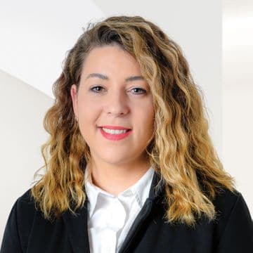 Portrait of a woman with wavy light-colored hair wearing a dark blazer and white shirt, facing the camera. The photograph is set against a light neutral background.