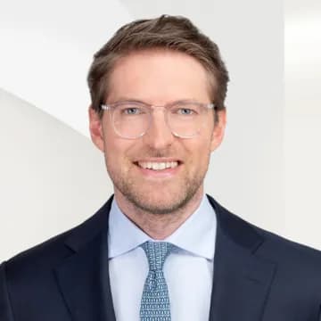 Gerrit Hunsdick mit hellbraunen Haaren und einer klaren, runden Brille, der einen marineblauen Anzug und eine blau gemusterte Krawatte trägt, vor einem hellen geometrischen Hintergrund.