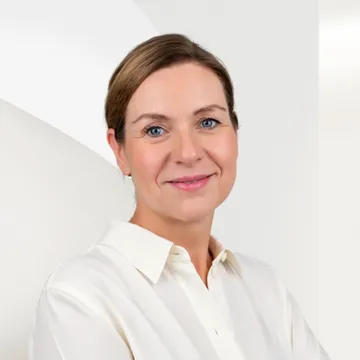Lächelnde Anke Fortkamp mit kurzen hellbraunen Haaren, die ein weißes Hemd trägt und vor einem blassen geometrischen Hintergrund posiert.