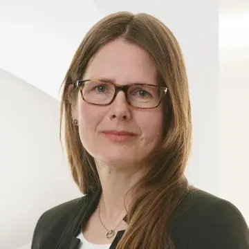 A woman with long brown hair and glasses, wearing a black blazer and white top, stands against a light background.